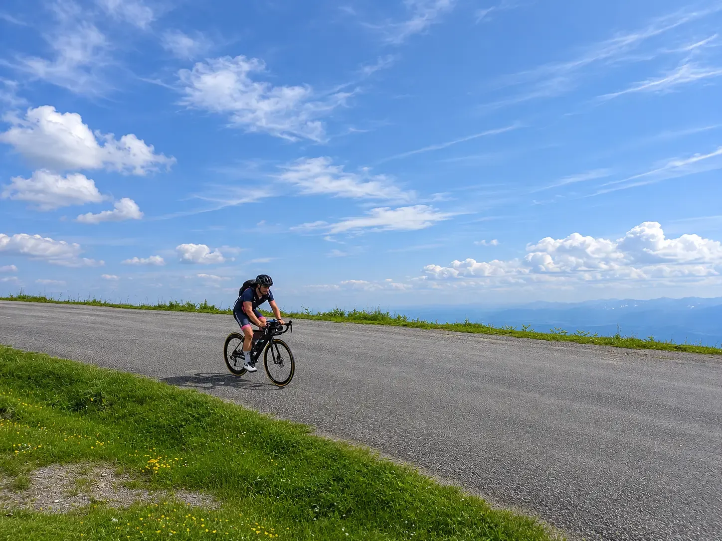 Un cycliste roule sur une route de montagne ensoleillée, sous un vaste ciel bleu parsemé de nuages blancs avec des reliefs lointains à l'horizon.